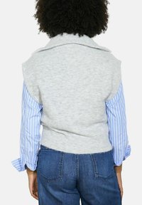 Cilvēks ar cirtainiem matiem, ģērbies gaismas pelēkā bezpiedurkņu adītā veste pār zilo un balto svītraino krekli un ziliem denimu džinsiem, redzams no aizmugures.