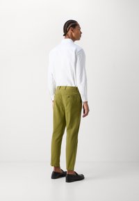 Chemise blanche légère et pantalon fuselé vert olive, avec une texture lisse et des poches arrière avec un détail de bouton. Souliers noirs à enfiler.
