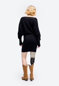 Kobieta o blond włosach, ubrana w czarną sukienkę i brązowe buty, idąca od nas, z widoczną protezą prawej nogi poniżej kolana.