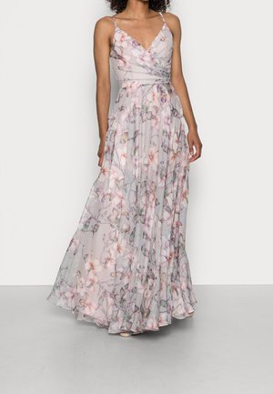 Femme portant une robe longue au sol, sans manches, rose pâle à fleurs, avec un décolleté en V et une jupe fluide.