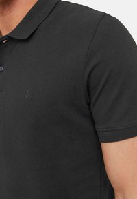 Polo shirt nera in tessuto texturizzato, con tre bottoni al colletto e un logo discreto sul petto. Maniche corte e design aderente.