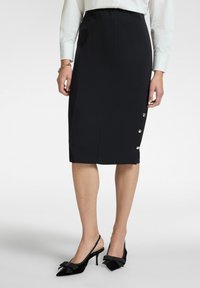 Zwarte pencilrok van stretchstof met een gladde textuur en zijknopen. Gecombineerd met zwarte slingback hakken met strikaccenten.
