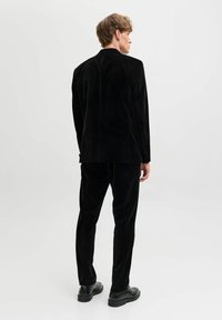 Čierny sametový oblek, úzky strih. Dlhý sako s klapkovými reverzami a rovné nohavice. Textúrovaná tkanina, minimálne označenie, v kombinácii s čiernymi topánkami.