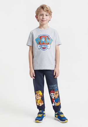 Jeune garçon debout portant un t-shirt Paw Patrol, un pantalon foncé avec les personnages de Paw Patrol, et des baskets bleues et jaunes sur fond blanc.