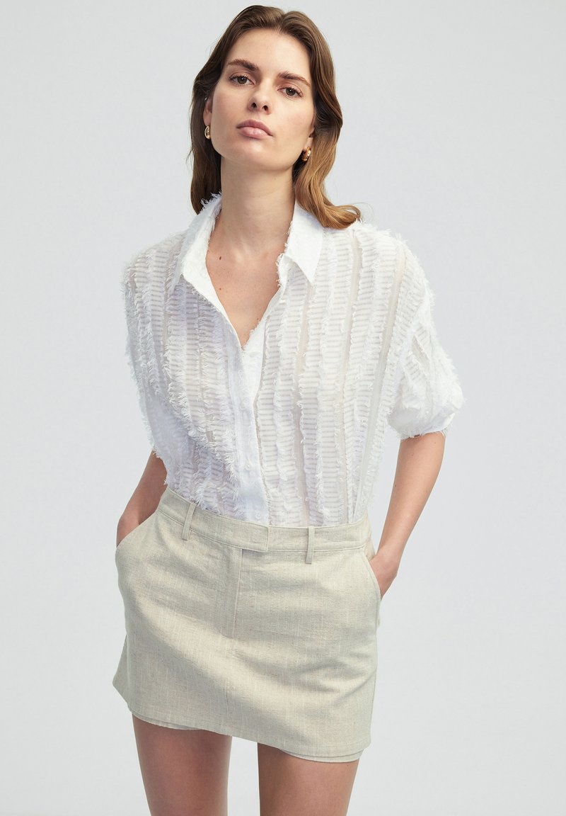 Chemise blanche texturée à manches courtes avec un devant à boutons et un col, associée à une mini-jupe taillée beige clair avec des poches.