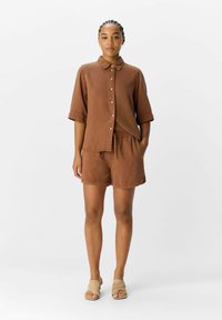 Camicia a maniche corte in lino marrone con bottoni e pantaloni short coordinati. L'outfit presenta una vestibilità comoda e tasche laterali, abbinato a sandali color nocciola.