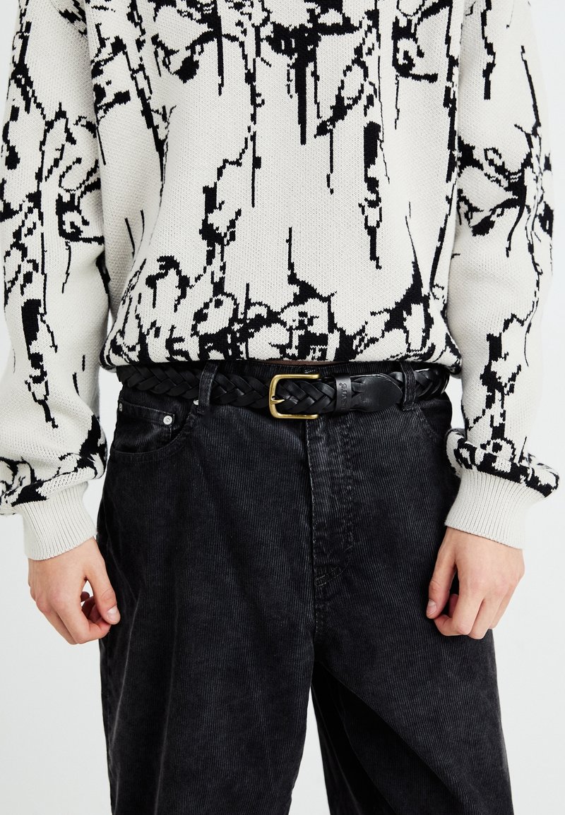 Personne portant un pull à motif abstrait blanc et noir rentré dans un pantalon en velours côtelé noir avec une ceinture en cuir tressé noire à boucle dorée.