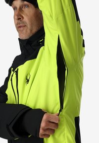 Gelb-schwarz Outdoor-Jacke mit Reißverschlüssen und einem inneren Netzfutter, das ein Spinnenlogo auf der Brust sowie eng anliegende Bündchen aufweist.