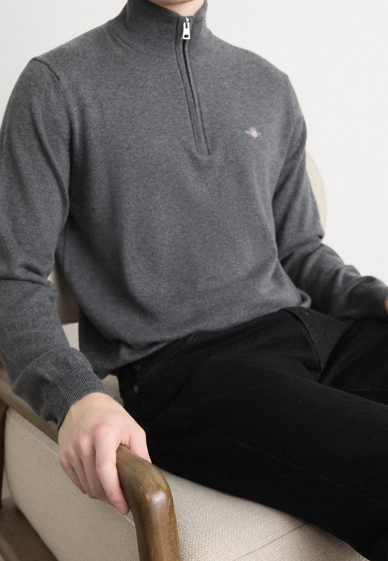 Personne portant un pull zippé gris et un pantalon noir, assise sur une chaise beige, tenant un accoudoir en bois avec la main gauche.