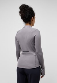 Top sportivo grigio chiaro a maniche lunghe con un design aderente, tessuto testurizzato, aperture per i pollici e un dettaglio riflettente sulla schiena.
