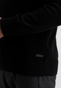 Zwarte gebreide trui met geribbelde manchetten en zoom. Heeft een klein metalen label aan de zijkant. De textuur lijkt zacht en van gemiddeld gewicht te zijn.