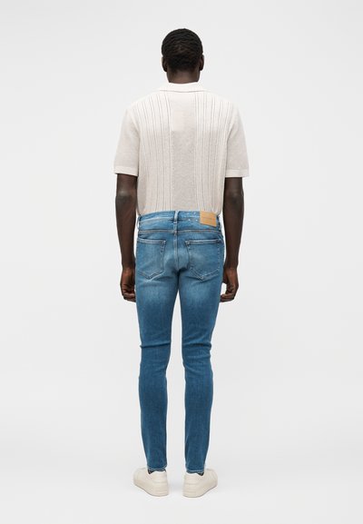 Άνδρας που είναι γυρισμένος με την πλάτη φοράει κρεμ πλεκτό κοντομάνικο πουκάμισο, στενά μπλε τζιν και λευκά αθλητικά παπούτσια σε απλό ανοιχτό φόντο.