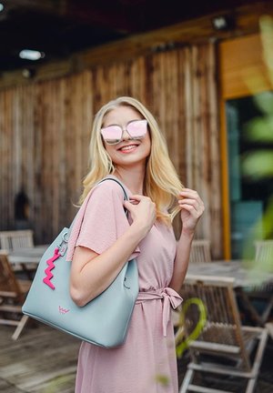 Gray handbag with a pink zig-zag charm; smooth texture, streamlined design. Model wears a light pink dress and mirrored sunglasses.