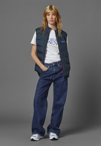 Marinblå pufferväst över vit T-shirt med tryckt text, matchad med löst sittande blå jeans och grå sneakers på en neutral bakgrund.