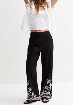Femme portant un pantalon noir à jambes larges avec une broderie florale blanche à l'ourlet, un haut blanc sans manches et une chemise courte à manches ouvertes.