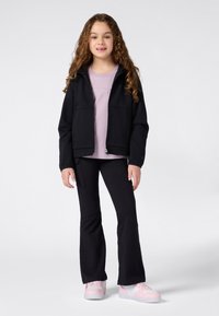 Ung jente med langt krøllete hår iført en svart zip-up jakke, lys lilla t-skjorte, svarte bukser og rosa-hvite joggesko som står foran en ensfarget bakgrunn.