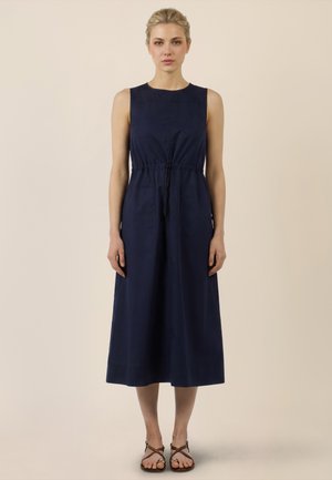 Blonde Frau steht, trägt ein ärmelloses, marineblaues Midikleid mit Taillenband und braune, flache Riemchensandalen, neutraler beiger Hintergrund.