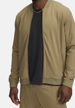 Man wearing beige zip-up jacket over black shirt, beige pants, and silver chain necklace, standing against white background.