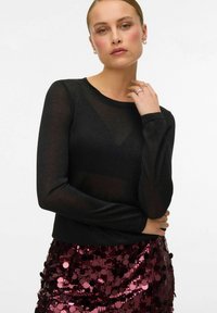 Czarna bluzka z długim rękawem z przezroczystym materiałowym nakładką, zestawiona z błyszczącą, bordową spódnicą z cekinami w kształcie okręgów.