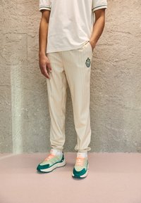 Person som bär krämfärgade rutiga byxor med en lapp, vit T-shirt och gröna och persikofärgade sneakers, stående mot en strukturerad vägg.