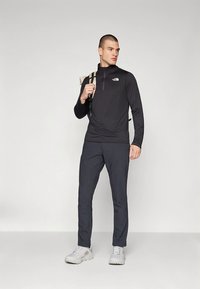 Must pikkade varrukatega lukuga särk The North Face logoga, kombineeritud tumedate pükste ja hallide jalanõudega; modell hoiab seljakotti.