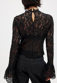 Schwarzes Spitzenoberteil mit langen Ärmeln und ausladenden Manschetten, Knopfverschluss am Hals hinten und einem aufwendigen floralem Muster mit Punktakzenten.