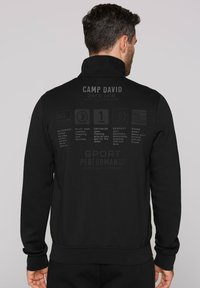 Zwarte sweatshirt met hoge kraag, met grafische tekst op de achterkant. Materiaal lijkt zacht, met een standaard pasvorm en geribbelde manchetten.