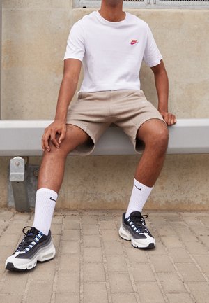 Osoba siedzi na metalowej ławce, ubrana w białą koszulkę Nike, beżowe szorty, białe skarpetki Nike oraz czarno-białe buty sportowe Nike.