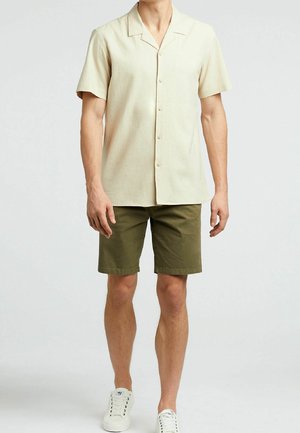 Mężczyzna w kremowej, krótkiej koszuli na guziki, oliwkowych szortach i białych trampkach stoi na jednolitym tle.