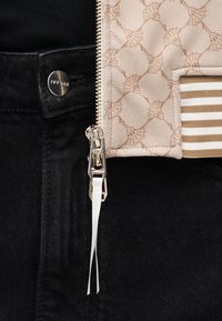 Κοντινή λήψη μαύρων τζιν με μεταλλικό κουμπί με την επιγραφή IVY OAK και ασημένιο φερμουάρ σε μπεζ υφασμάτινο φόντο με ριγωτό μανίκι.