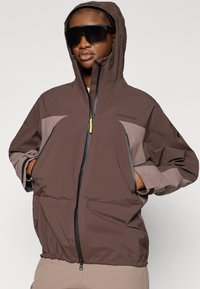 Αδιάβροχο μπουφάν με κουκούλα σε σκούρο καφέ χρώμα με ανοιχτό καφέ λεπτομέρειες· διαθέτει κλείσιμο με φερμουάρ, ελαστικές μανσέτες και τσέπες με φερμουάρ.