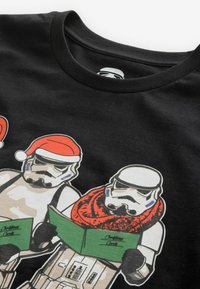 Černé bavlněné tričko s dvěma figurkami stormtrooperů, jeden v červeném šálu a vánoční čepici, oba drží zelené vánoční pohlednice s textem.