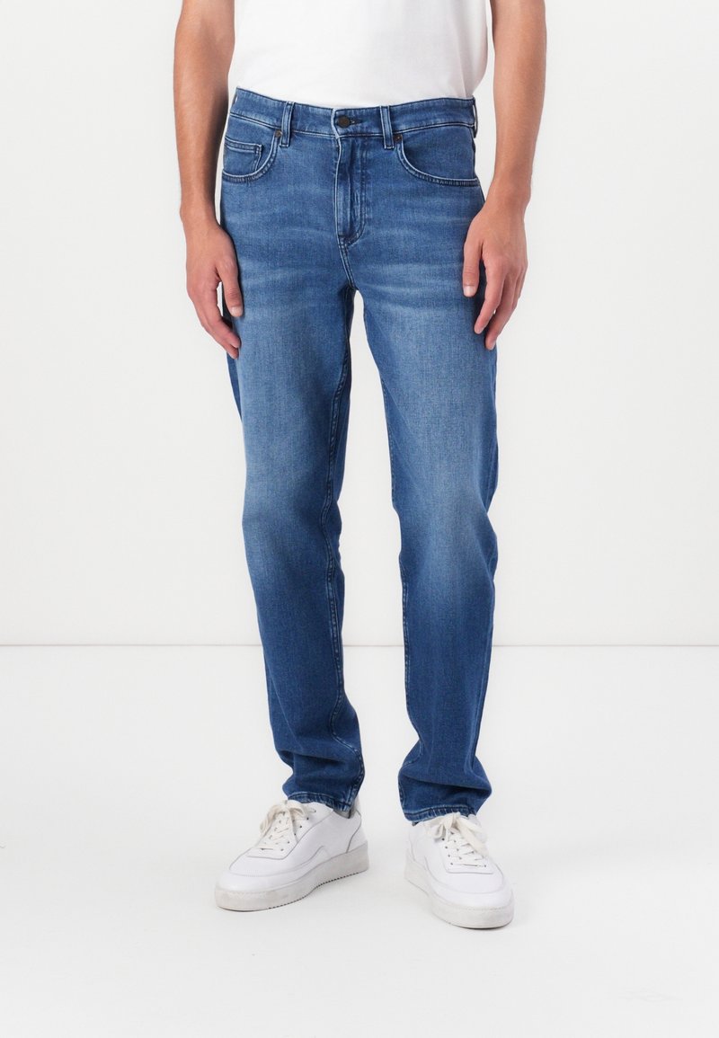 Homme portant un jean droit bleu et des baskets blanches, debout devant un fond blanc uni, les mains détendues le long du corps.