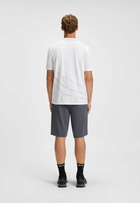 Hvid kortærmet atletisk skjorte med et gråt abstrakt design på ryggen, parret med mørkegrå shorts og sorte løbesko.
