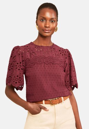 Frau trägt eine burgunderfarbene Lochspitzenbluse mit Puffärmeln und einem braunen Gürtel und steht mit einer Hand in der Tasche hellbeiger Hose.