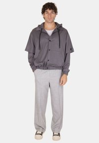 Giacca grigia a maniche corte con cappuccio, cordoncini regolabili, bottoni frontali e due tasche, abbinata a pantaloni a pieghe grigi chiari e sneakers.