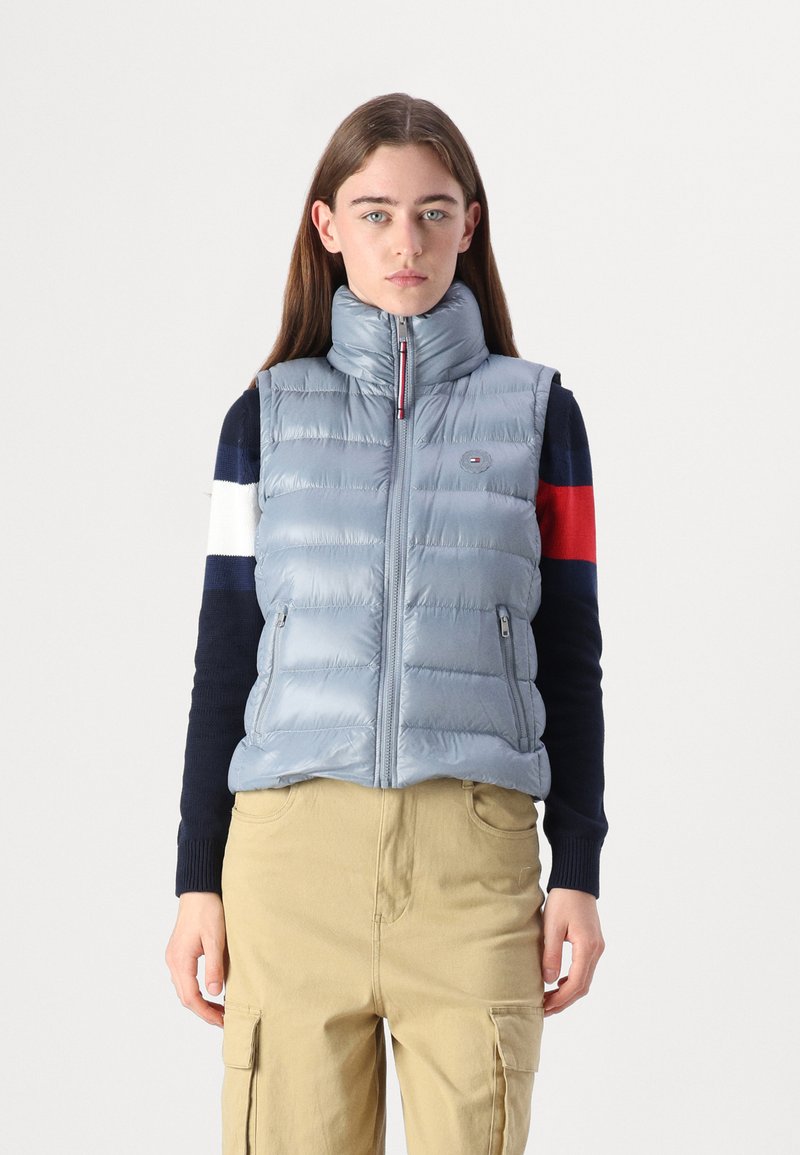Gilet rembourré bleu clair avec col montant, fermeture éclair à l'avant et deux poches latérales. Comprend des sections matelassées et un badge logo sur la poitrine.