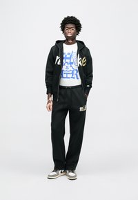 Čierna mikina s kapucňou Nike so zlatým nápisom, biele tričko s modrým grafickým dizajnom, čierne tepláky s nápisom "nike" a sivé tenisky.