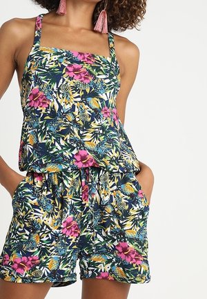 Femme portant une combinaison sans manches à motif floral avec des couleurs rose, bleu, jaune et vert, mains dans les poches, et boucles d'oreilles à pampilles roses.
