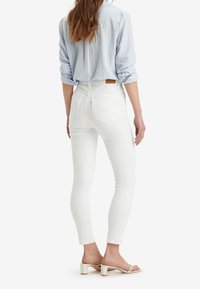 Mujer vestida con pantalones ajustados blancos de cintura alta, camiseta de rayas azul claro con las mangas remangadas y sandalias de tacón beige, de pie mirando hacia atrás.