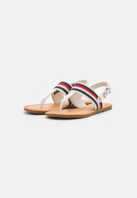 Un paio di sandali aperti con suole color ocra, cinturini bianchi e bande di tessuto rosse, bianche e blu navy sopra e cinturini a fibbia posteriori.