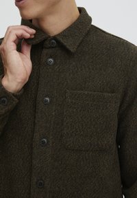 Chaqueta de camisa de lana verde oscuro con cierre de botones en el frente, cuello y un bolsillo en el pecho. La tela tiene una apariencia texturada y ligeramente moteada.