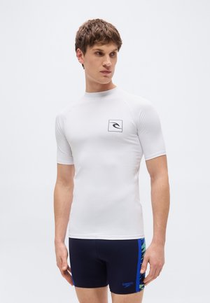 Νεαρός άνδρας που φοράει λευκή κοντομάνικη μπλούζα προστασίας από τον ήλιο και σκούρο μπλε μαγιό με μπλε και πράσινες πλαϊνές λωρίδες, στέκεται μπροστά σε απλό φόντο.