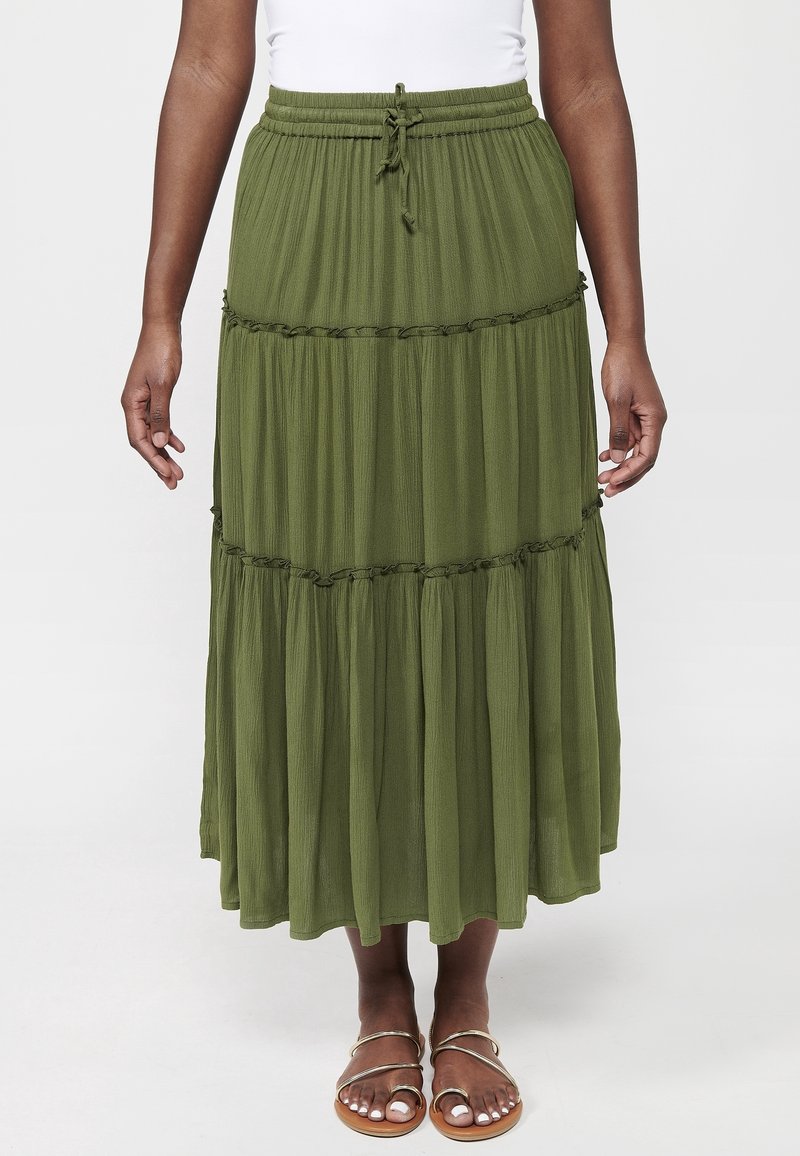 Personne portant une longue jupe à volants vert olive avec une taille à cordon et des sandales à brides métalliques, debout devant un fond uni.