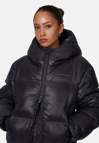 Giacca a vento nera con colletto alto e cappuccio oversize. Realizzata in materiale lucido e liscio con cuciture orizzontali e zipper frontale.