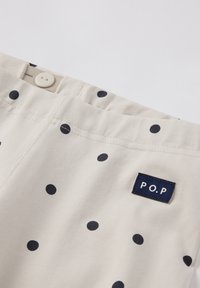 Beige-kangas shortsit, joissa on laivastonsinisiä pilkkuja, varustettuna nappisulkimella ja pienellä laivastonsinisellä tarralla, jossa lukee "P.O.P.", ommeltuna vyötärölle.