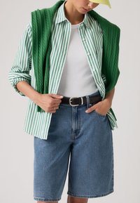 Άτομο που φοράει πουκάμισο με πράσινες και λευκές ρίγες, λευκό αμάνικο, πράσινο πουλόβερ στους ώμους, τζιν σορτς, μαύρη ζώνη και κίτρινο καπέλο.