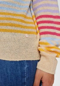 Detailní záběr na ruku oblečenou v pleteném svetru s vícebarevnými pruhy v žluté, modré, fialové a růžové barvě, který je přes modré džíny.