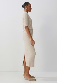 Beiges Strickkleid mit kurzen Ärmeln und einem seitlichen Schlitz, aus texturiertem Stoff. Kombiniert mit Riemchensandalen, seitlich dargestellt.