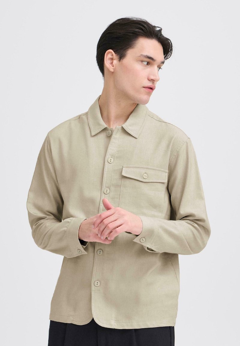 Svetlobéžová košeľa na gombíky s golierom, jedným náprsným vreckom a dlhými rukávmi s gombíkovými manžetami. Hladká tkanina a voľný strih.